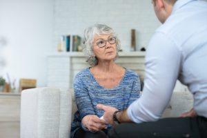 Elderly woman in a blue sweater having a serious conversation with a healthcare professional, illustrating the significance of 'Primary Health Care' in providing personalised support and advice. Background includes a cosy, well-lit living room setting.