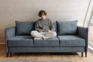 A young man with curly hair sits cross-legged on a blue sofa, using a smartphone. The setting suggests a calm, indoor environment, potentially indicating the use of technology for remote mental health assessments.