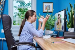 A young woman sits at a desk, holding a tissue and gesturing while on a video call with a therapist displayed on her computer screen. The background features plants and a bright blue wall, highlighting the use of technology for mental health assessments.