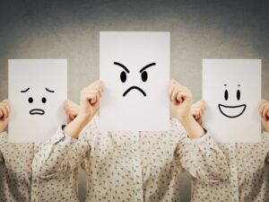 Three people holding papers with happy, angry, and worried faces, representing emotions in an NDIS therapy session.