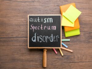 Blackboard with "Autism Spectrum Disorder" written in chalk. Sticky notes and coloured chalk pieces on a wooden table. Causes of speech delay include autism.